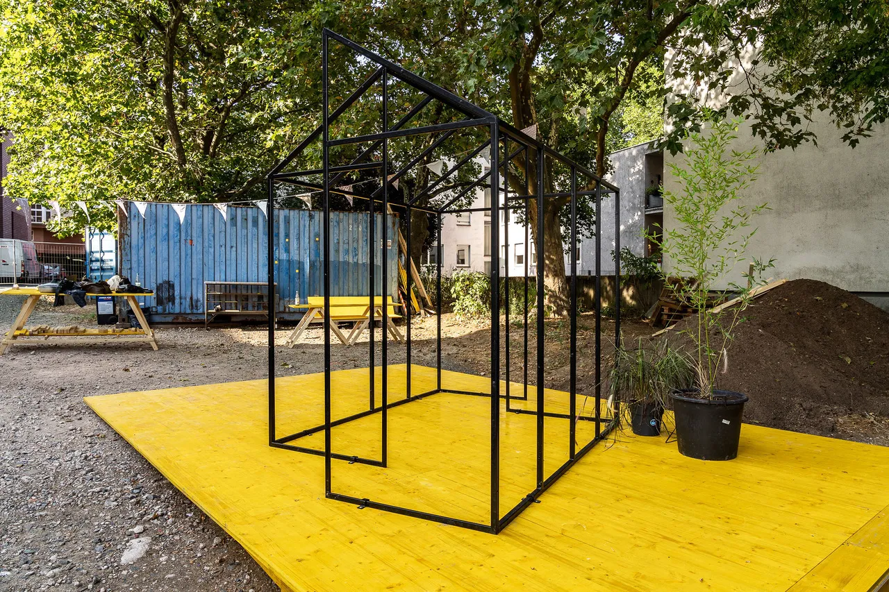 Ein stilisiertes Haus als Metall-Rahmen-Modell auf einer gelben Fläche