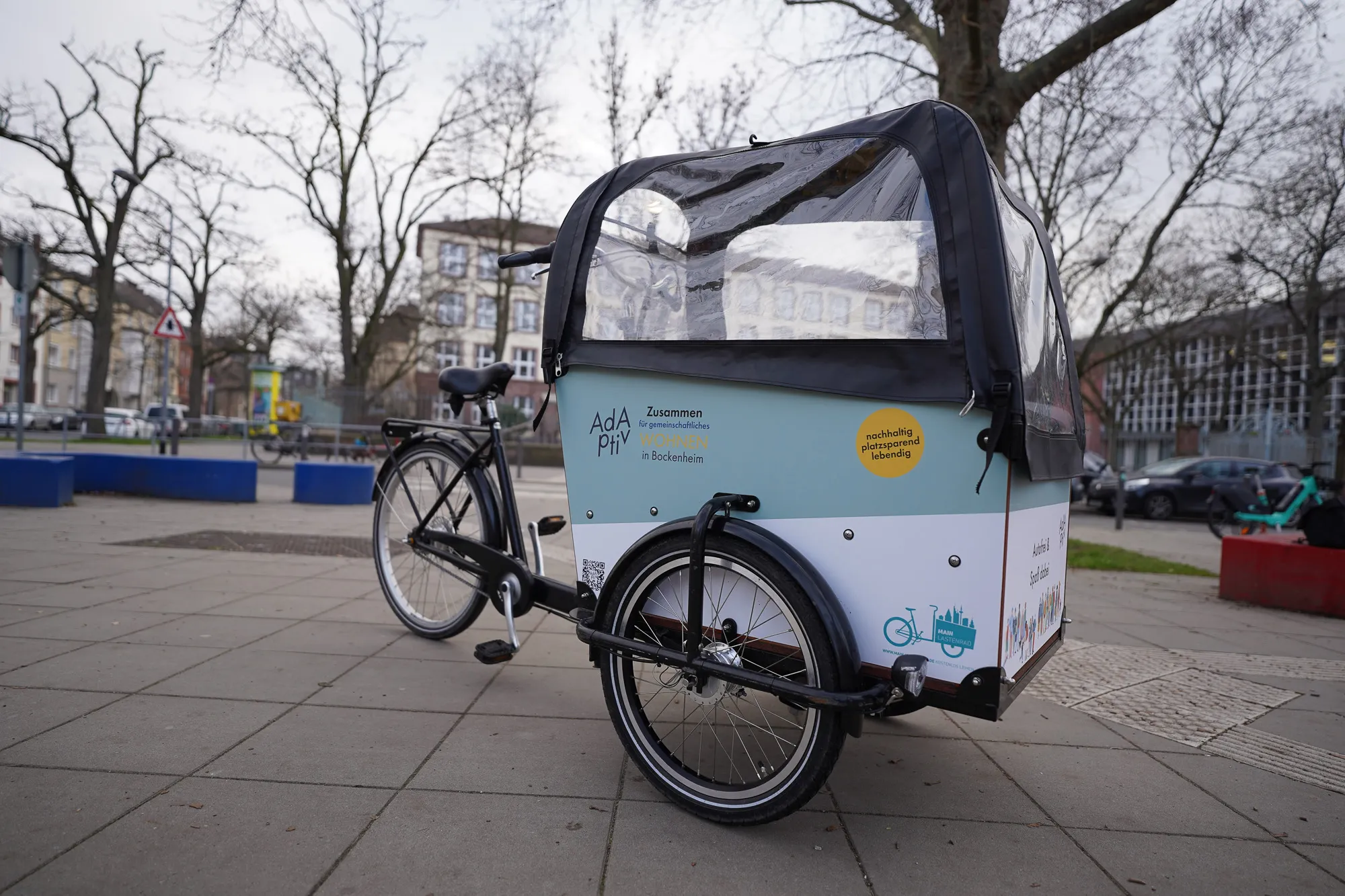 Ein dreirädriges Lastenrad mit großer Ladefläche und Regenverdeck. Die Seitenwände sind mit dem Logo von AdAptiv und Informationen über das Projekt bedruckt.