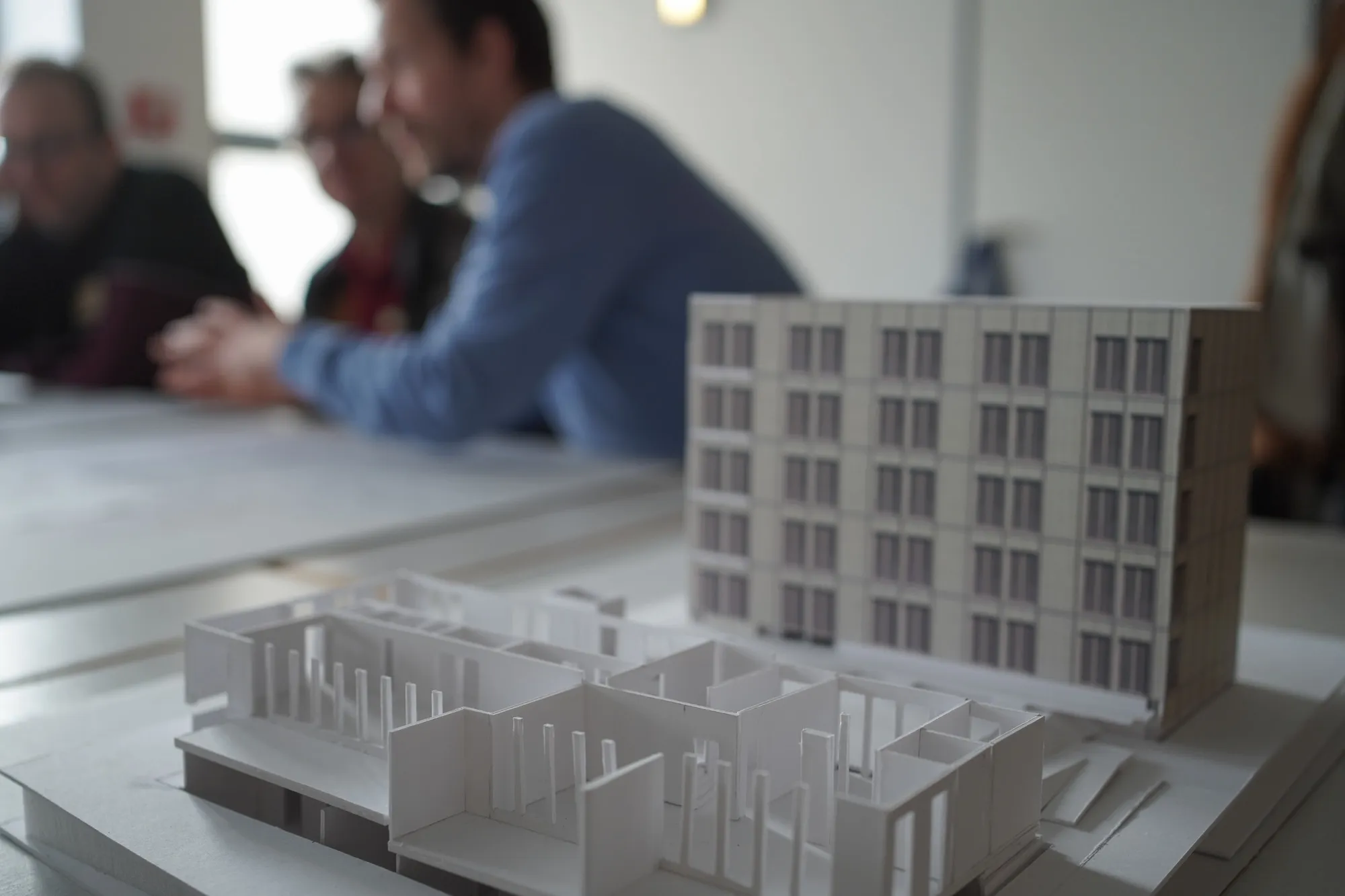 Im Vordergrund ein Architektur-Modell, eine Gebäudehälfte hat keine Fassade und zeigt die Innenwände des Ergeschosses. Im Hintergrund unscharf Menschen die über einen Grundriss diskutieren.